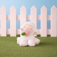 Plush toy sheep with a pink bubble gum sitting on grass with a white picket fence in the background