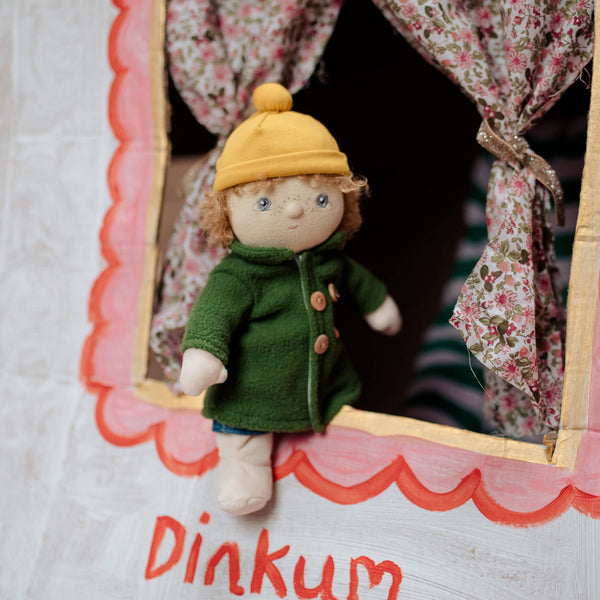 Small doll in a green coat and yellow hat peeking out from behind floral curtains with 'Dinkum' written below.