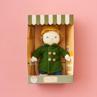 Stuffed doll in a green coat with a yellow hat in a box on a pink background